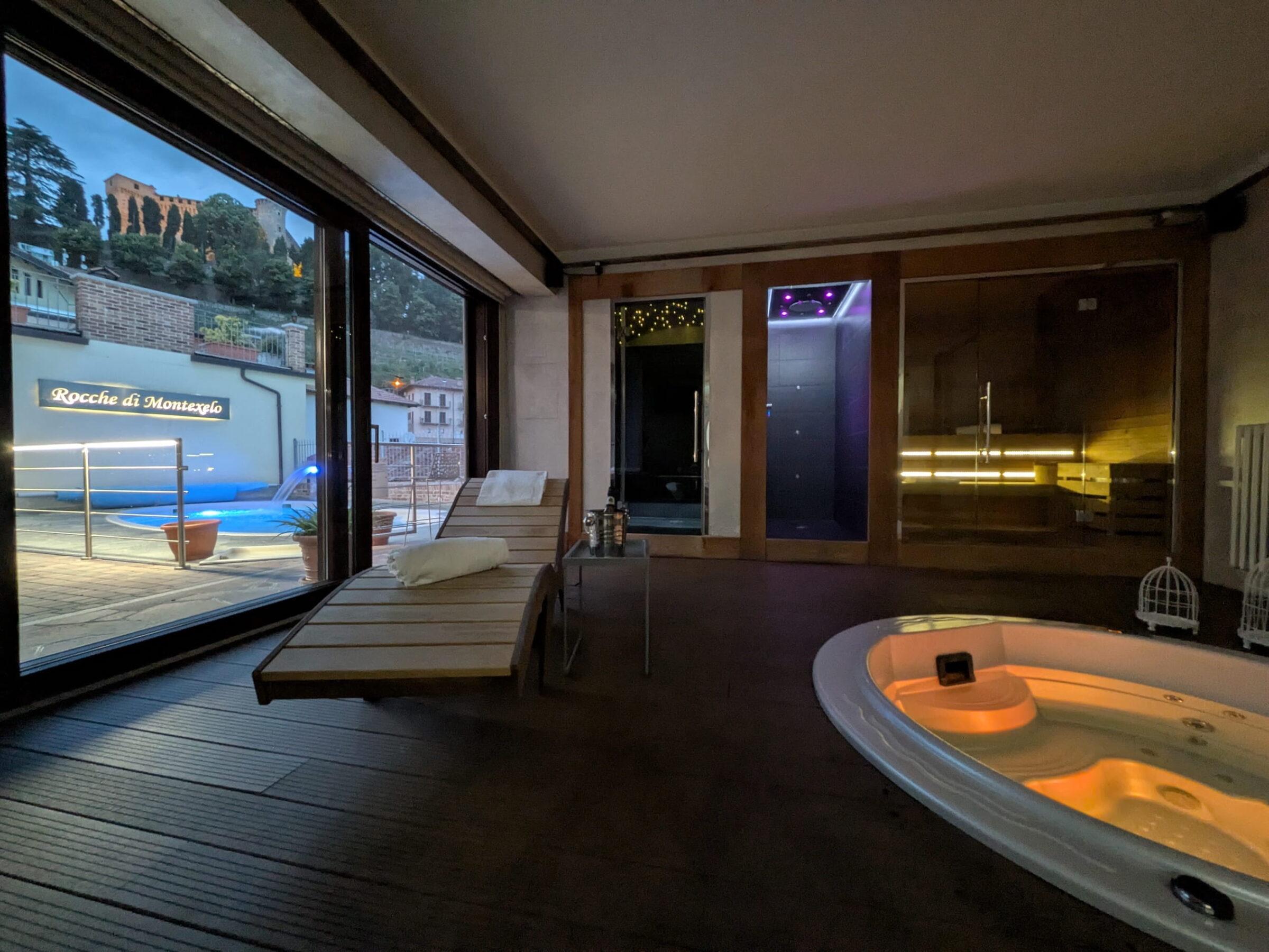 Wellnessbereich im Rocche di Montexelo mit einem beleuchteten Whirlpool, zwei Liegestühlen, einem großen Fenster mit Blick auf die Terrasse und den Hügel mit seinen Ruinen sowie sanfter Beleuchtung