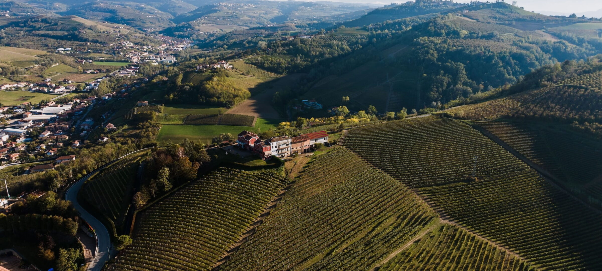 Luftaufnahme von hügeliger Landschaft mit Weinbergen und vereinzelten Gebäuden in der Region Piemont