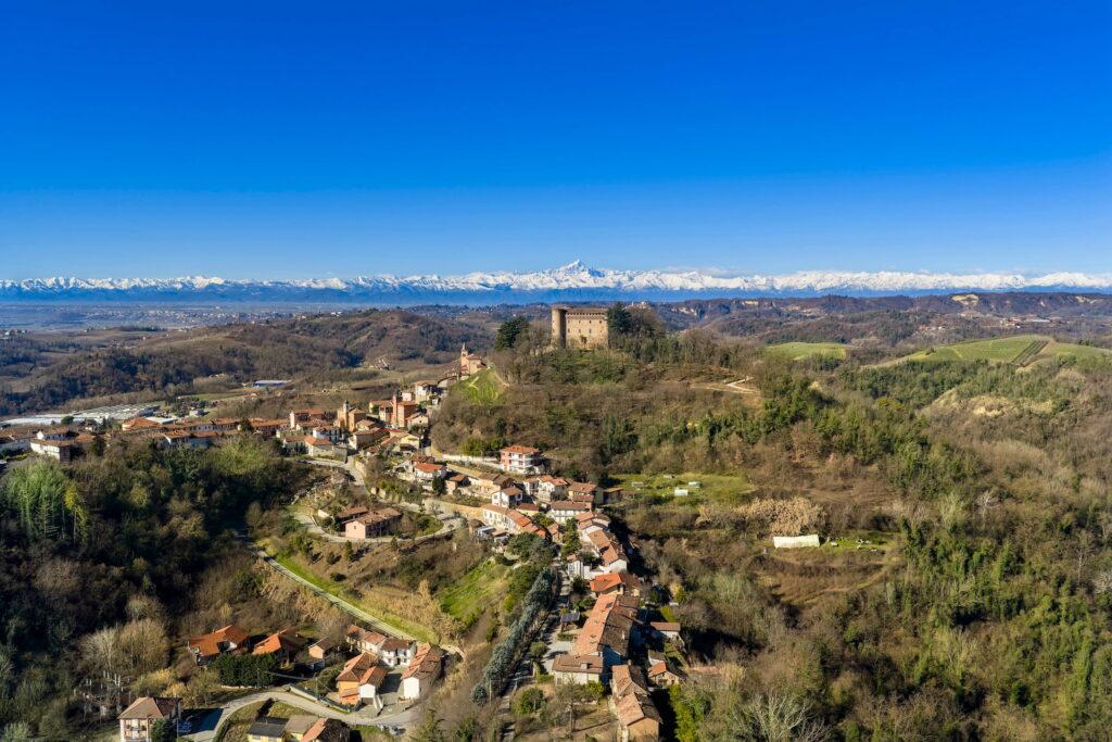 Panoramic view of Monticello d'Alba, featuring a castle on a wooded hill with snow-capped mountains in the background in the Piedmont region