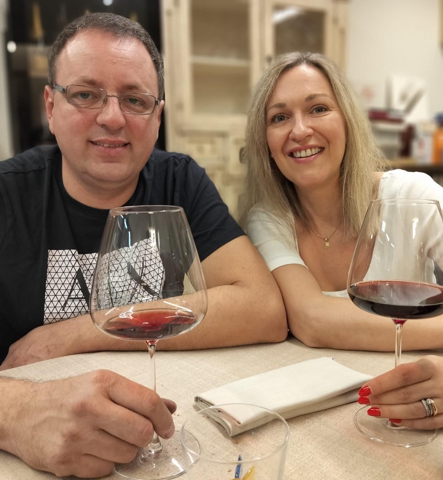 Two smiling people are sitting at a table, each holding a glass of red wine; they are the owners of Rocche di Montexelo in Piedmont